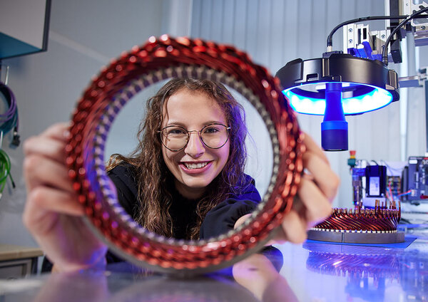 Eine junge Frau lächelt, während sie einen Stator in der Hand hält, umgeben von moderner Technik.