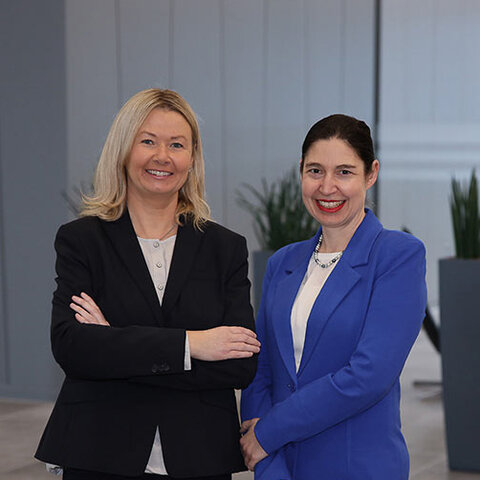Zwei Frauen stehen in einem modernen Büro, lächeln und strahlen Selbstbewusstsein aus, umgeben von eleganter Architektur.