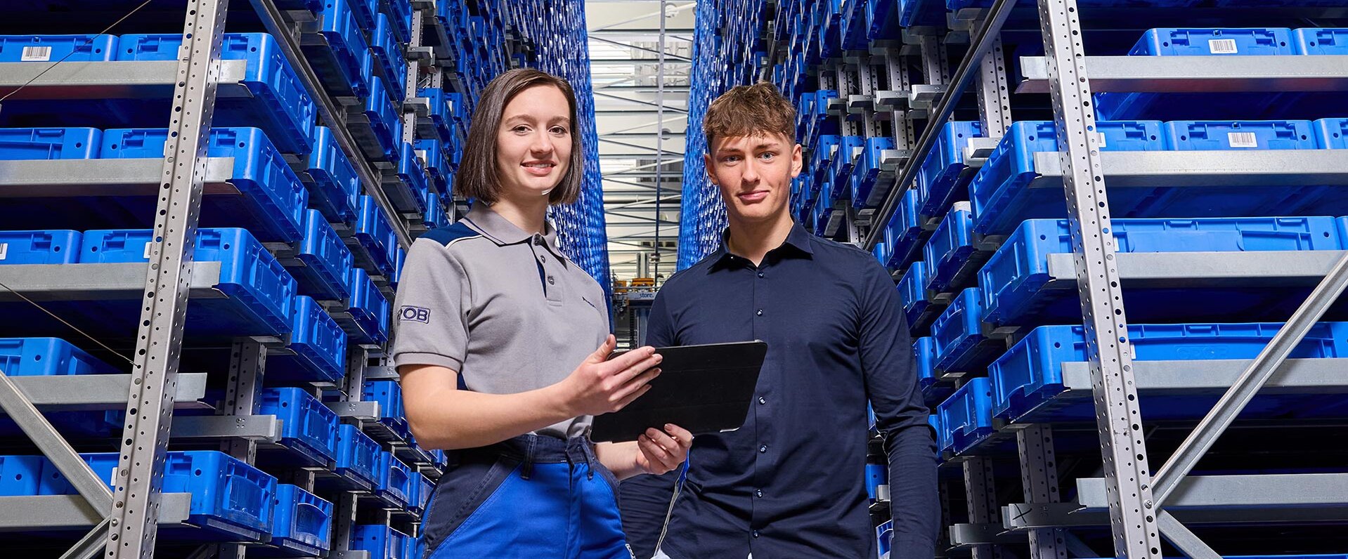 Die Mitarbeiter von GROB stehen in einem gut organisierten Lager, das mit blauen Regalen gefüllt ist, und nutzen ein Tablet zur Kommunikation.