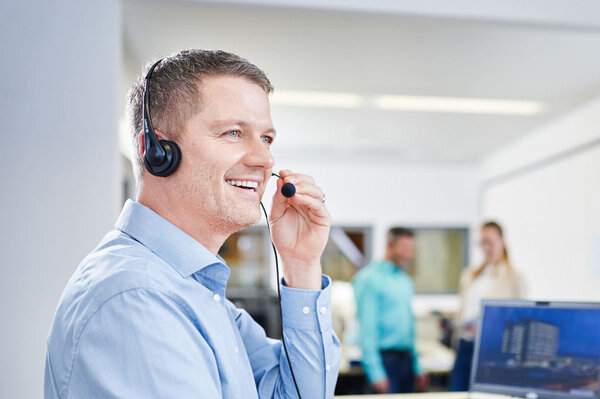 Ein lächelnder Mitarbeiter von GROB trägt ein Headset und spricht aktiv in ein Mikrofon, während er an einem Schreibtisch sitzt. Im Hintergrund sind zwei Kollegen zu sehen, die sich unterhalten, was eine freundliche und kooperative Arbeitsatmosphäre vermittelt.