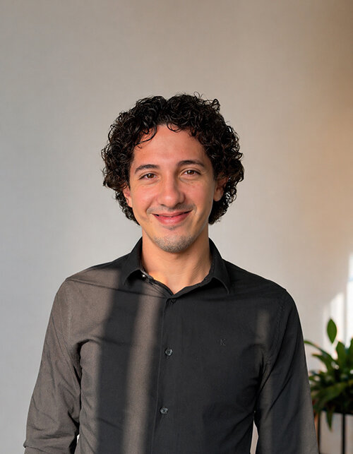 Ein junger Mann mit lockigem Haar lächelt freundlich vor einem hellen Hintergrund, der eine positive Atmosphäre ausstrahlt.