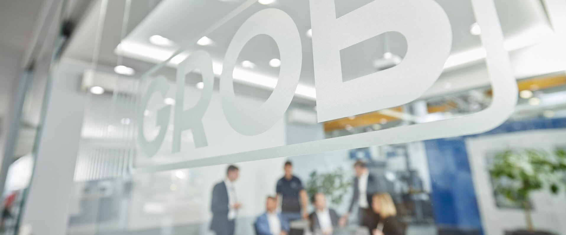 Die Glaswand mit dem GROB-Logo im Vordergrund zeigt eine moderne Büroumgebung mit Mitarbeitern in einem Meeting.