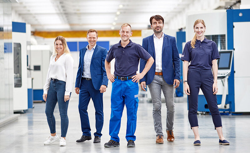 Fünf Mitarbeiter von GROB stehen in einer modernen Produktionshalle, lächelnd und in Arbeitskleidung, bereit für neue Herausforderungen.