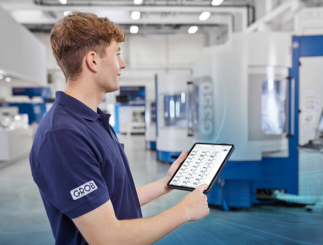 Ein Mitarbeiter von GROB steht in einer modernen Fertigungshalle und nutzt ein Tablet zur Überwachung von Maschinen.