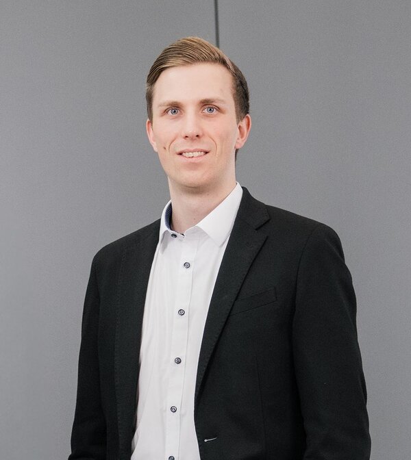 Florian Grob in einem eleganten Anzug steht an einer Rolltreppe und lächelt selbstbewusst in die Kamera.