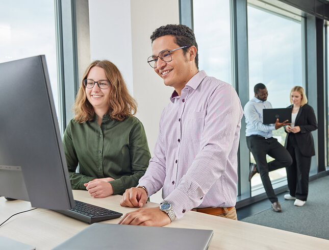 Ein freundliches Team von GROB diskutiert am Arbeitsplatz, während sie an einem Computer arbeiten und sich gegenseitig unterstützen.