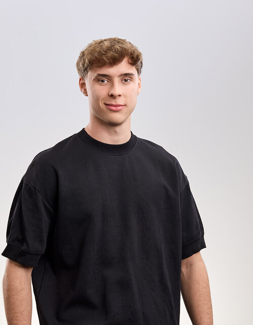 Ein junger Mann mit lockigem Haar trägt ein schwarzes T-Shirt und lächelt selbstbewusst in die Kamera.
