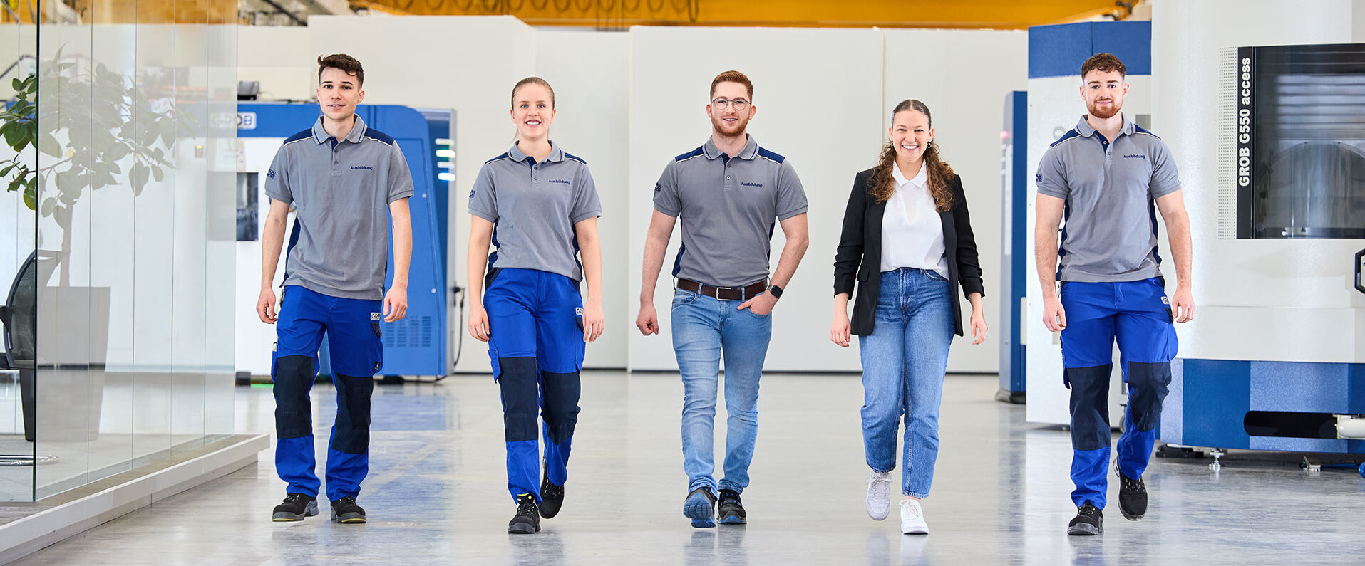 Die Gruppe von GROB-Auszubildenden zeigt Teamgeist, während sie in Arbeitskleidung durch die lichtdurchflutete Fabrikhalle schreitet.