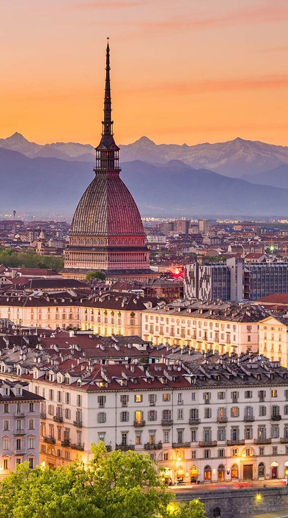 Die Skyline von Turin bei Sonnenuntergang, mit dem markanten Mole Antonelliana im Vordergrund und den Alpen im Hintergrund.