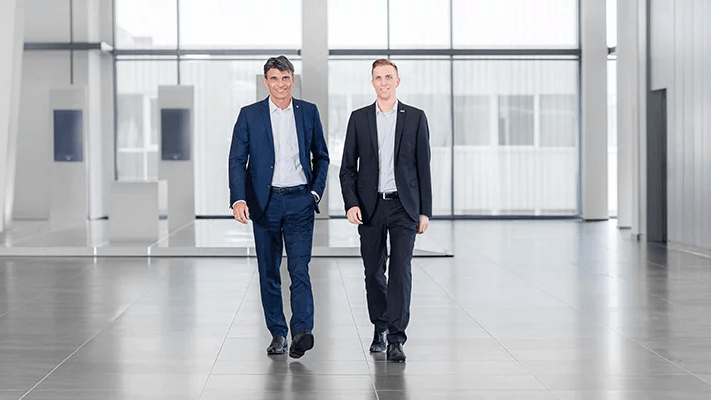 Zwei professionell gekleidete Männer bewegen sich durch einen stilvollen, hellen Raum, der durch große Fenster und klare Linien geprägt ist.