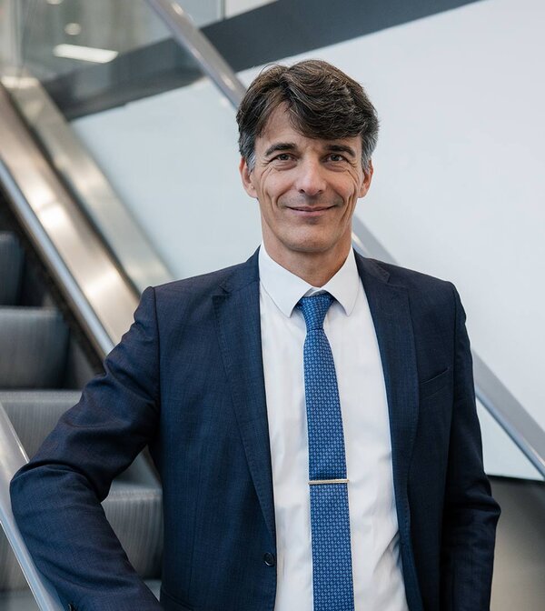 Aufsichtsratsvorsitzender Christian Grob steht an einer Treppe, gekleidet in einem eleganten Anzug und einer blauen Krawatte.