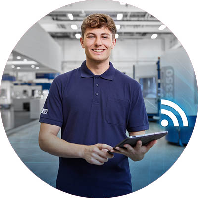  Ein junger Mann in einem blauen Poloshirt lächelt, während er ein Tablet in einer modernen Industrieumgebung hält.